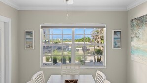 A dining area with a large window offering a view of the beach and greenery. There are white chairs and a wooden table.