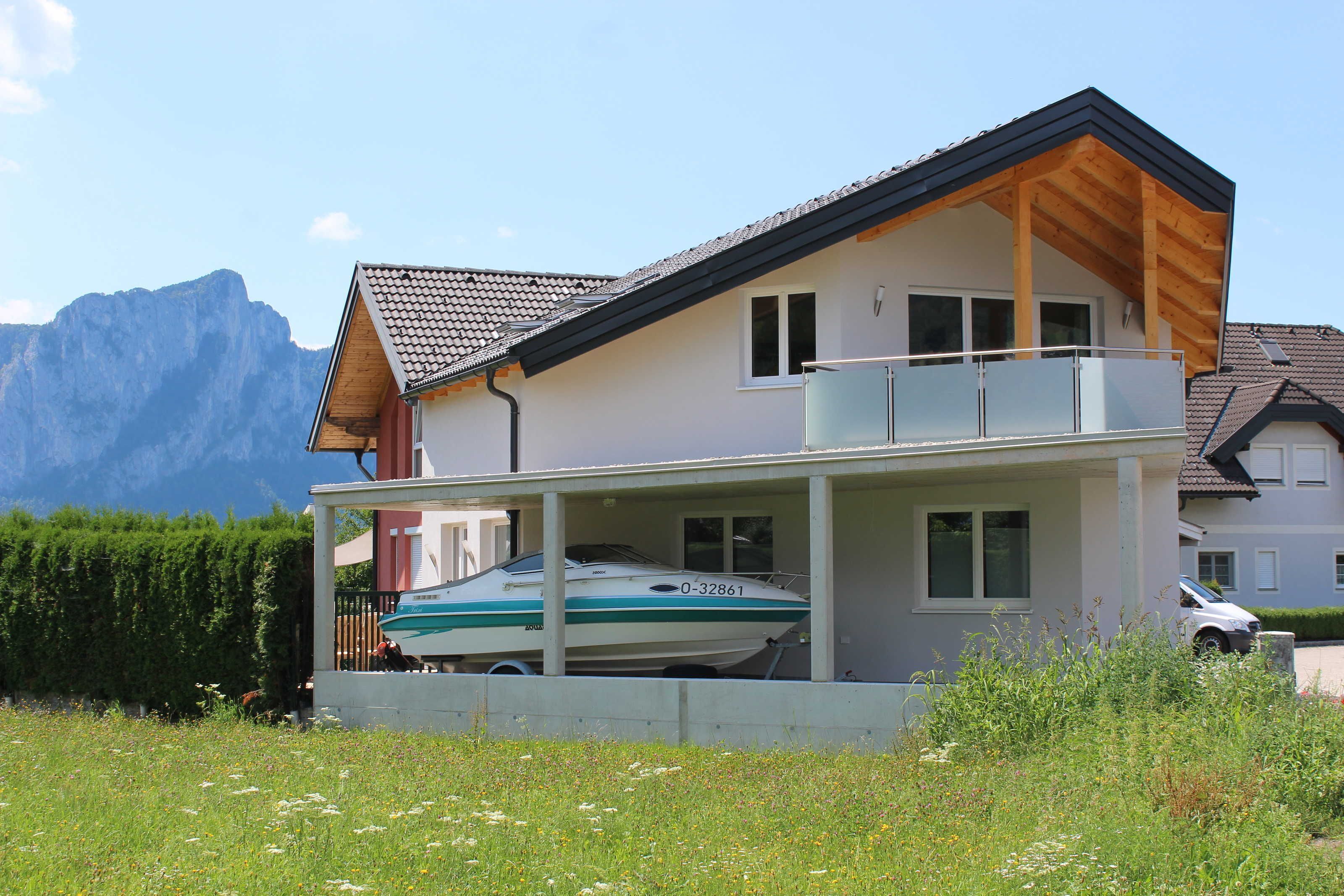 Ein zweigeschossiges Haus mit Balkon, einem Boot auf der Auffahrt und Bergen im Hintergrund.