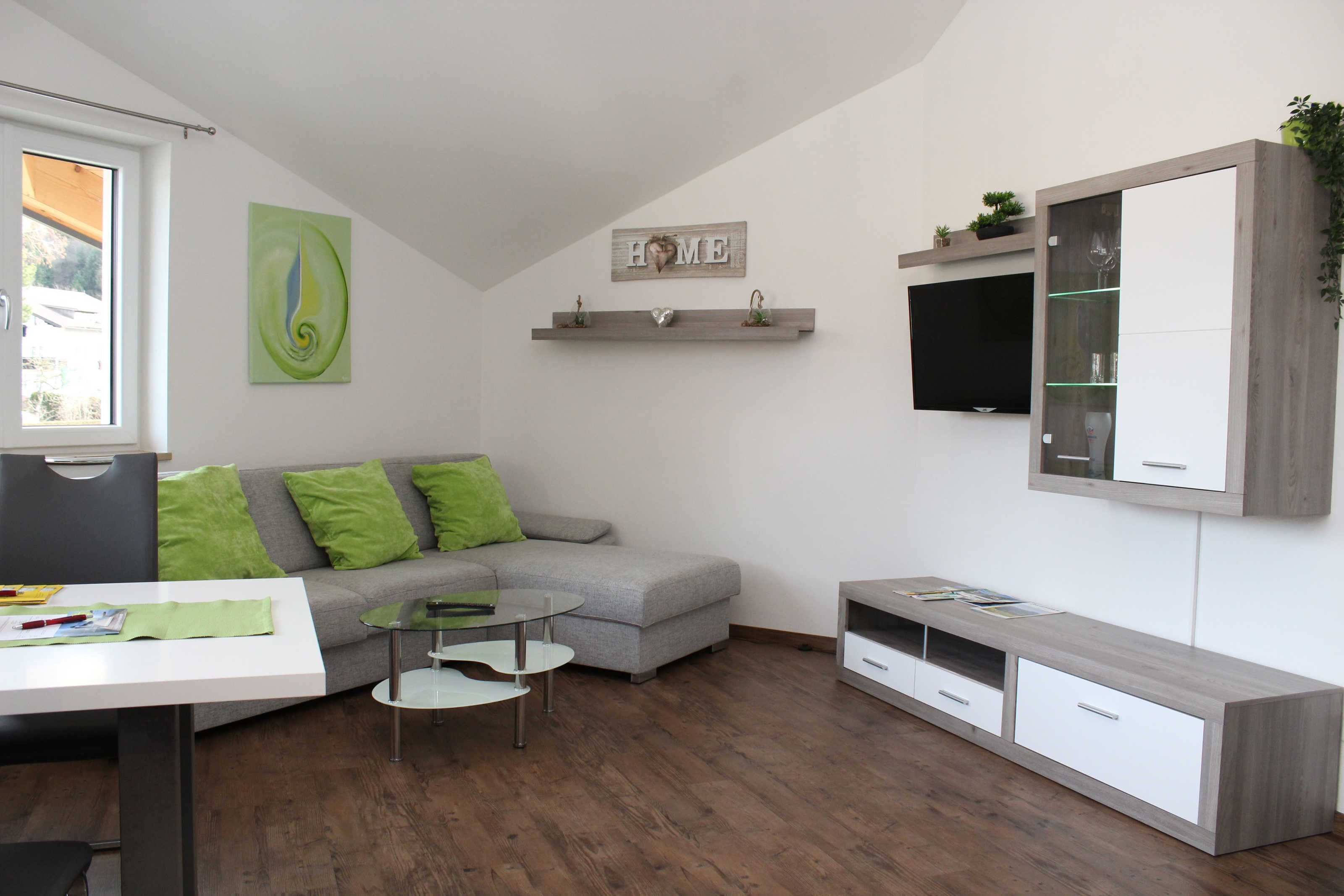 Ein modernes Wohnzimmer mit einem grauen Sofa, grünen Kissen, einem Glastisch, einer weißen TV-Konsole und einem Wandregal mit Dekoration.