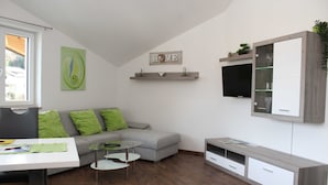 A modern living room with a grey sofa, green cushions, a glass coffee table, a white TV stand, and a mounted shelf with decorative items. - Apartment Lindpointner (Innerschwand am Mondsee)