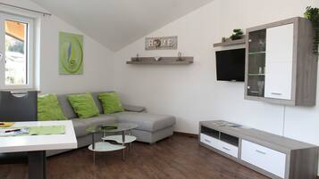 Ein modernes Wohnzimmer mit einem grauen Sofa, grünen Kissen, einem Glastisch, einer weißen TV-Konsole und einem Wandregal mit Dekoration.