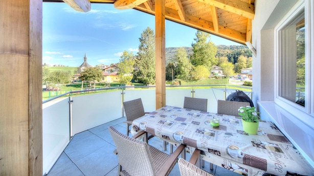 A covered outdoor patio with a table set for dining, wicker chairs, and a view of a village. - Apartment Lindpointner (Innerschwand am Mondsee)