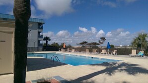Zona de piscina con sillones de descanso y sombrillas, un edificio con balcones y un cielo despejado.
