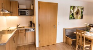 Une cuisine compacte équipée d’un four à micro-ondes, d’une machine à café et d’un lave-vaisselle. On y trouve une porte en bois, un banc et une table à manger avec des chaises.