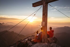 Une croix en bois, sur laquelle est assise une personne, surplombe un paysage montagneux au coucher du soleil.