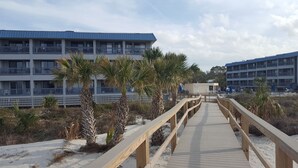 Una pasarela de madera que lleva a un edificio con balcones y una playa al fondo.