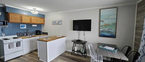 Una cocina compacta con armarios de madera, un refrigerador blanco, una zona de comedor con mesa y sillas, un televisor de pantalla plana montado en la pared, y un cuadro sobre el televisor.