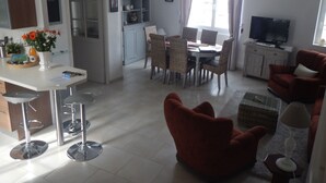 Ein modernes Wohnzimmer mit einem roten Sessel, einem Essbereich mit Korbstühlen, einer Küche mit weißen Schränken und einem Fernseher auf einem Schrank.