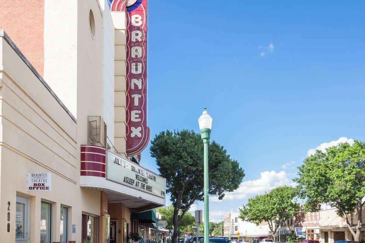 A theater building with a marquee reading 'BRAUNTEX' and a sign for 'JUL 3 BRAUNTEX THEATRE WELCOMES ASLEEP AT THE WHEEL.'