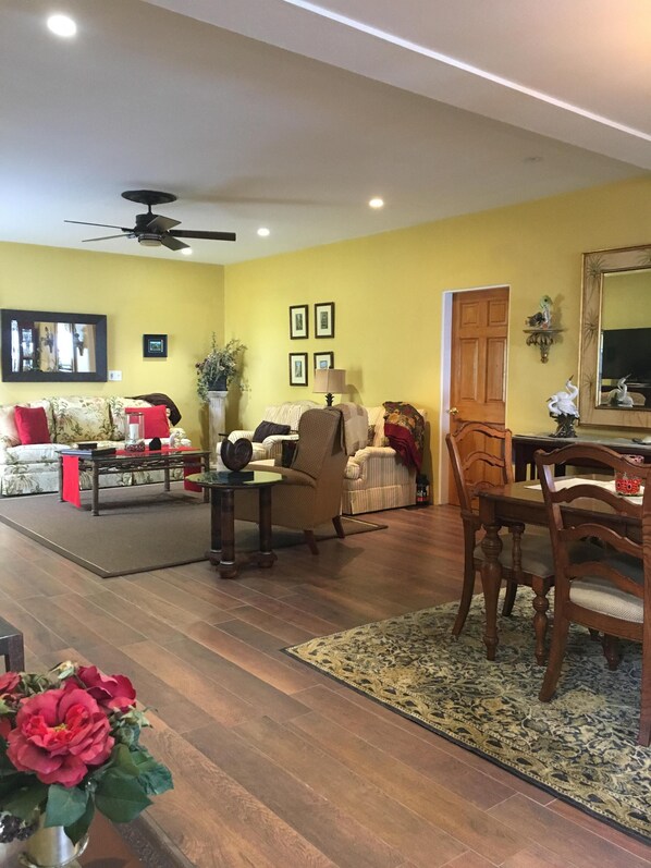 A living room with a sofa, armchairs, a coffee table, and a dining table with chairs. There are framed pictures on the wall, a ceiling fan, and a door leading to another room.