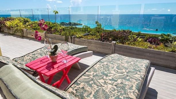 Una terraza con una mesa roja y cojines estampados de color verde, con vista a un océano azul y despejado.