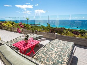 A deck with a red table and green patterned cushions, overlooking a clear blue ocean.