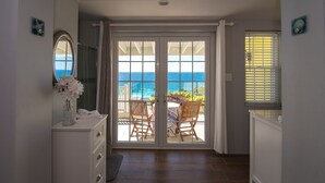 Habitación con puerta de vidrio corrediza que da a un balcón con vistas al océano. Se observa un aparador blanco con espejo y un florero, además de una pequeña mesa con sillas en el balcón.