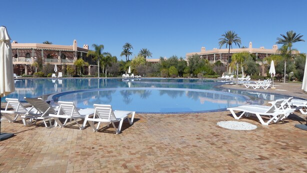 A pool area with white lounge chairs and umbrellas, surrounded by palm trees and a building with a tiled roof. - A haven of peace in the heart of the palm grove (Marrakech)