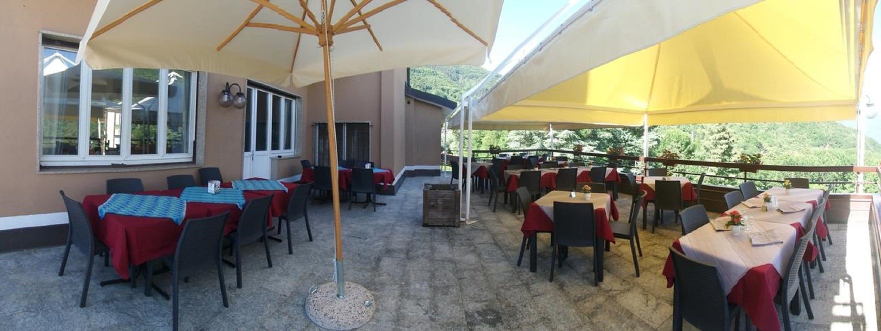 An outdoor patio area with tables and chairs, covered by large umbrellas, and a view of greenery in the background.