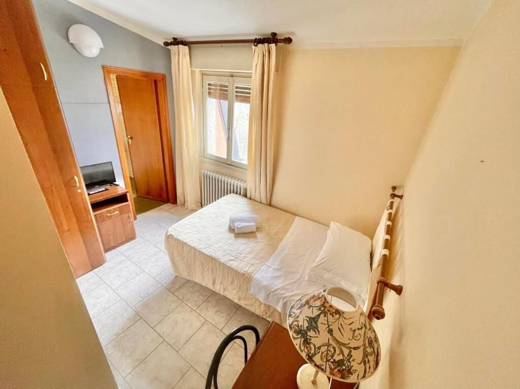 A bedroom with a bed, a bedside table, a lamp, a radiator, a window with curtains, and a door.