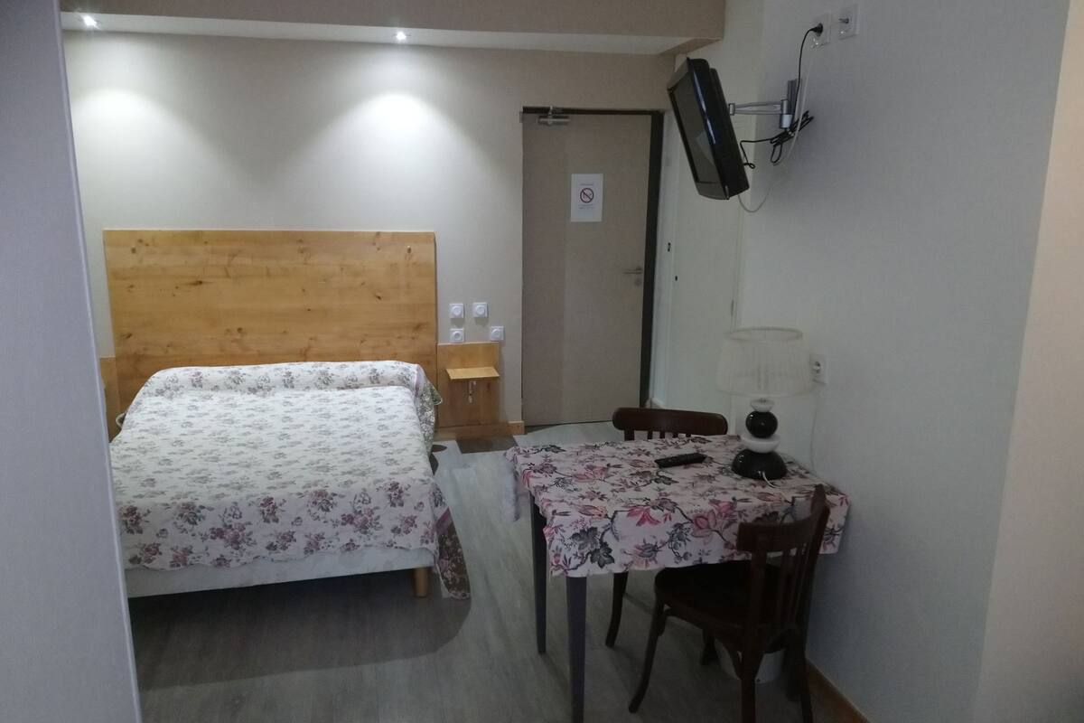 Une petite chambre propre, avec un lit doté d’une tête de lit en bois, un bureau avec une chaise, un téléviseur fixé au mur et une lampe.
