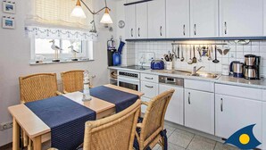 A modern kitchen with white cabinets, a dining area with wicker chairs, and a window with a seagull decoration. - Villa Strandperle, Whg. 22 - VS 22 (Heringsdorf)