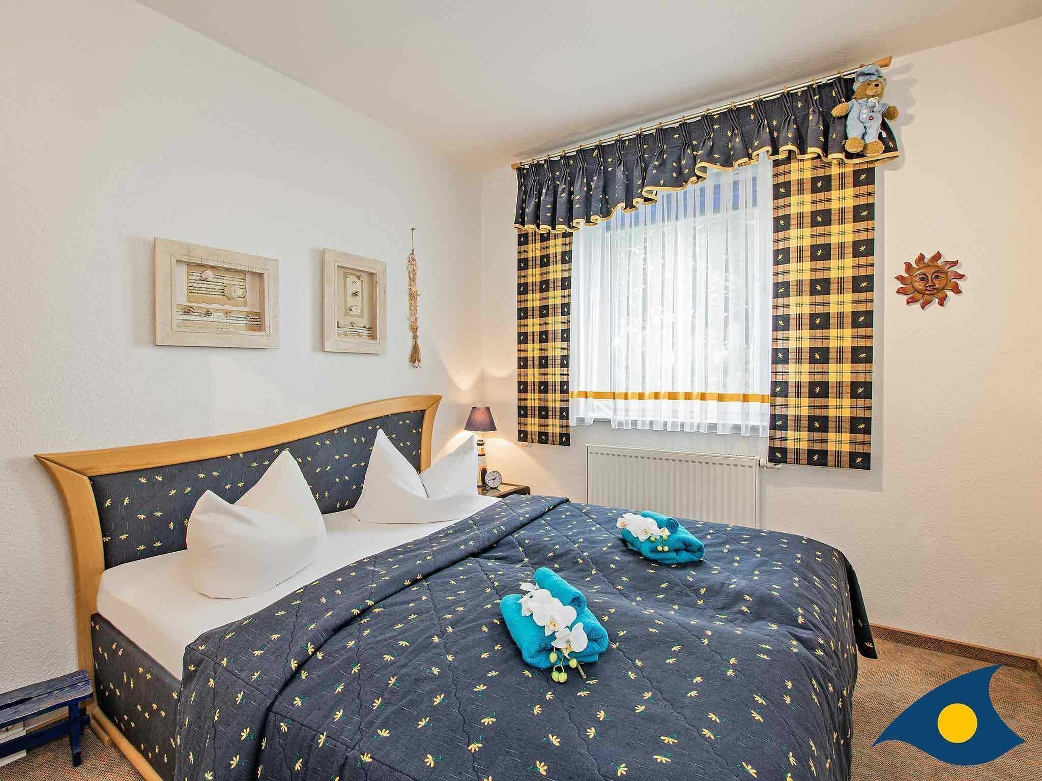 Una cama bien hecha con una colcha oscura estampada, almohadas blancas y una toalla azul. Hay dos cuadros en la pared, una ventana con cortina a cuadros y un pequeño adorno decorativo.