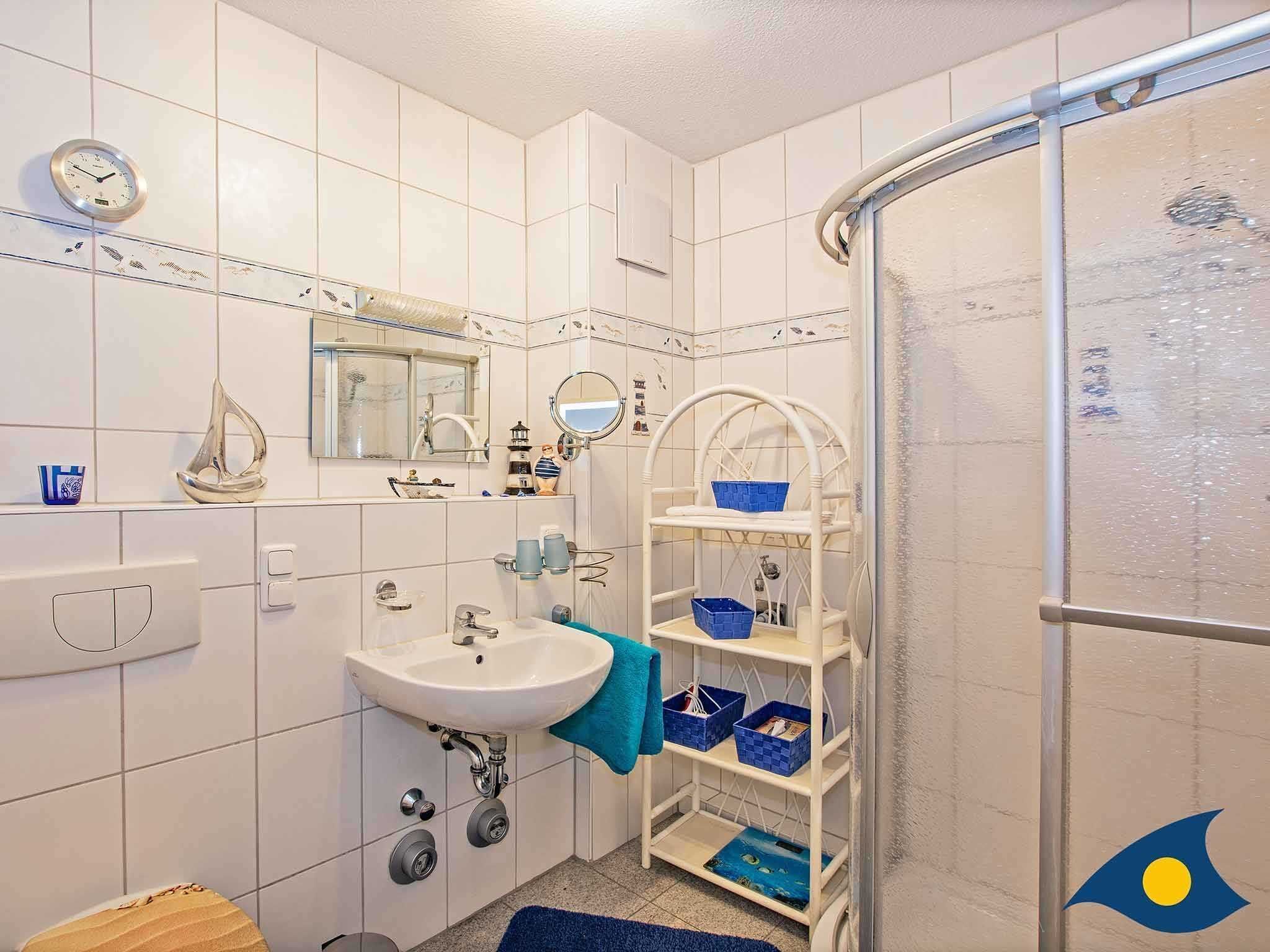 Un baño con lavamanos blanco, espejo, reloj y zona de ducha.