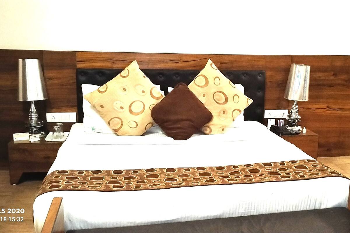 A hotel room with a wooden headboard, a bed with patterned pillows, two bedside tables with lamps, and a wooden chair.