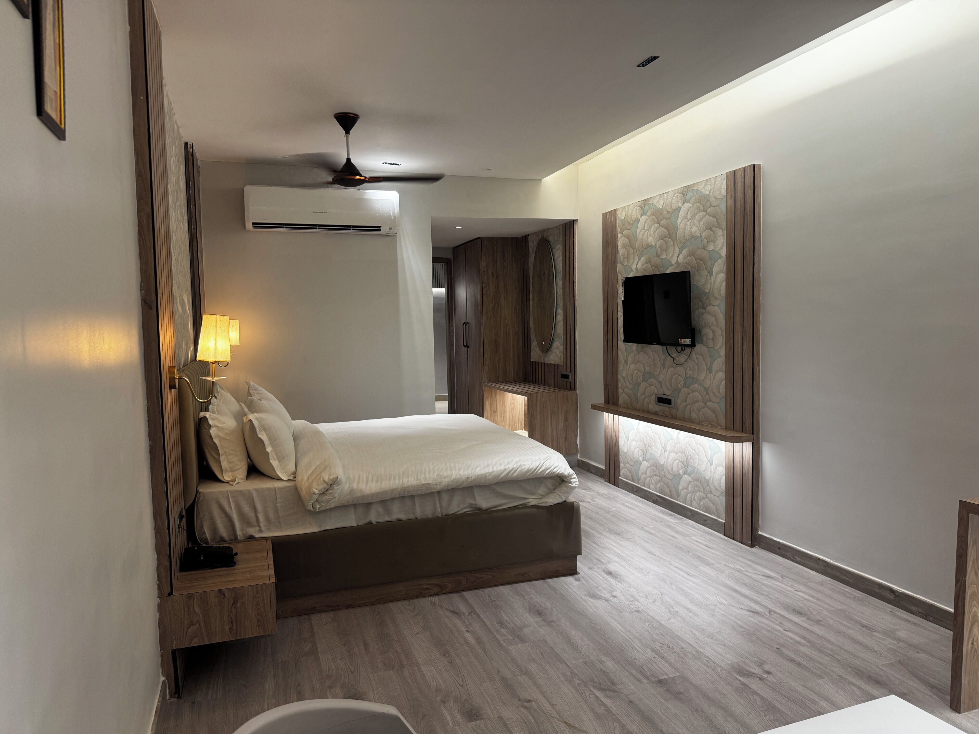 A modern bedroom with a bed, bedside lamp, air conditioning unit, ceiling fan, and a mounted TV.
