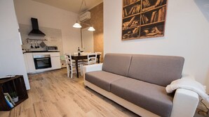 A modern living room with a grey sofa, a dining table, and a kitchen area with white cabinets and a stove.