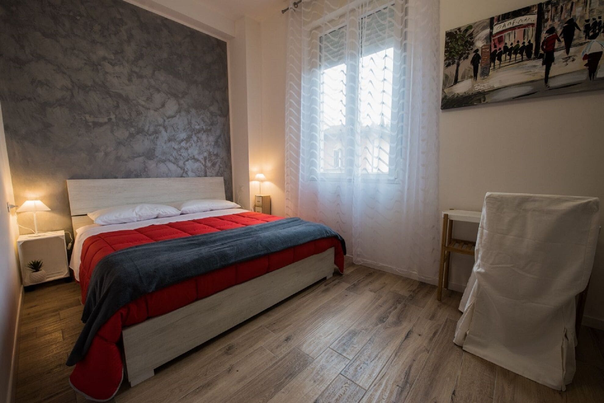 Un dormitorio con cama, mesitas de noche, una ventana con cortinas translúcidas y un cuadro en la pared.