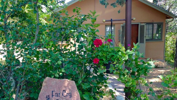 A stone sign with 'Far Cry Cottage' engraved, surrounded by greenery and rocks, leading to a house with a brown roof and a window with shutters.