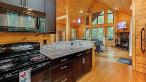A spacious log cabin with a kitchen featuring granite countertops, a stove, and dark wooden cabinets. There is a dining area with a table and chairs, and a fireplace.