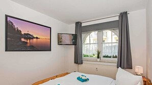 A hotel room with a bed, a window with curtains, a wall-mounted TV, and a framed picture of a sunset over water. - Villa Maria - Gabriele Whg, 02 - Maria 02 (Loddin)