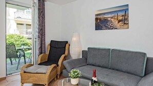 A living room with a grey sofa, wicker chairs, a glass coffee table with flowers and a bottle, and a beach view painting on the wall. - Villa Maria - Gabriele Whg, 02 - Maria 02 (Loddin)