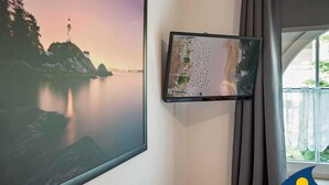 A room with a large framed photograph of a coastal landscape, a smaller monitor, and a window with curtains. - Villa Maria - Gabriele Whg, 02 - Maria 02 (Loddin)