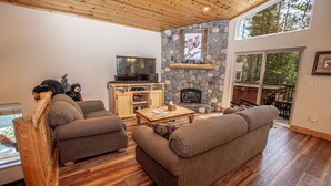 A cozy living room with a stone fireplace, wooden ceiling, and large windows overlooking a forest. - Sherman by Hauserman Rentals (Tahoma)