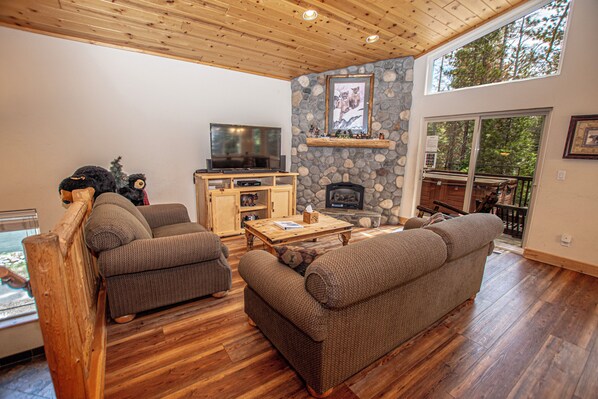 A cozy living room with a stone fireplace, wooden ceiling, and large windows overlooking a forest.