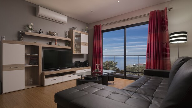 Una sala de estar moderna con un televisor de pantalla plana grande, un sofá de cuero y una mesa de comedor. Hay estantes con objetos decorativos y un amplio ventanal con cortinas.