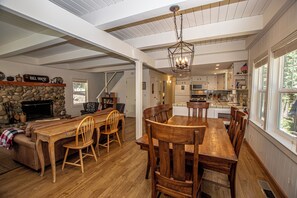 A spacious dining area with a wooden table and chairs, a stone fireplace, and a kitchen in the background. - Hillhouse by Hauserman Rental Group (Homewood)