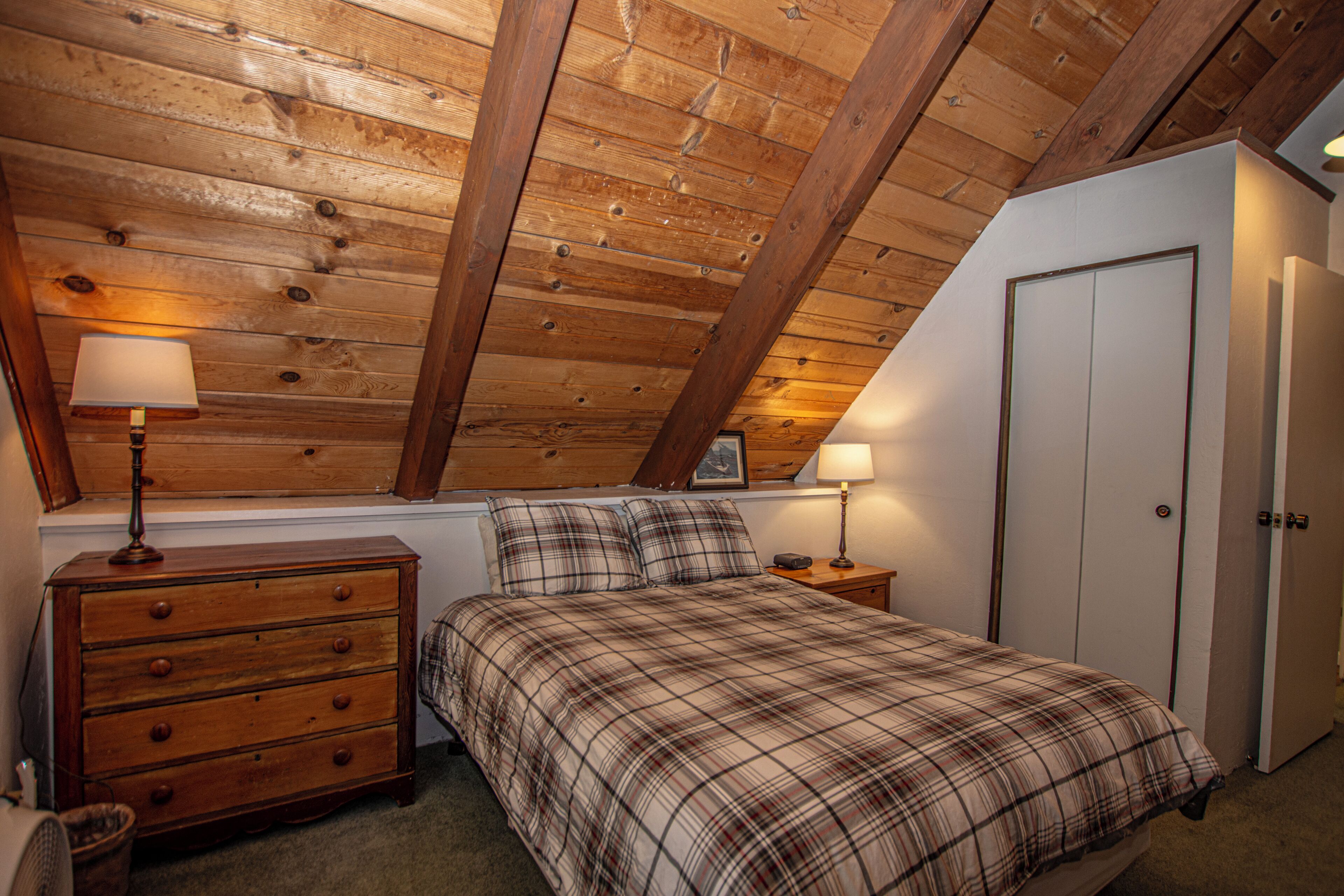 Ein Schlafzimmer mit Holzbalkendecke, einem Bett mit kariertem Überwurf, einer Kommode, einem Nachttisch mit Lampe und einer Tür.
