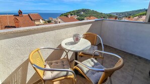 Een balkon met een ronde tafel en rieten stoelen, met uitzicht op een kustgebied met terracottedaken en groen.