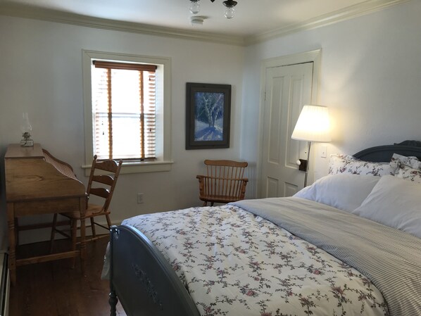 Une chambre à coucher comprenant un lit, un bureau, une chaise, une fenêtre avec des stores, un tableau encadré au mur et une table de chevet avec une lampe.