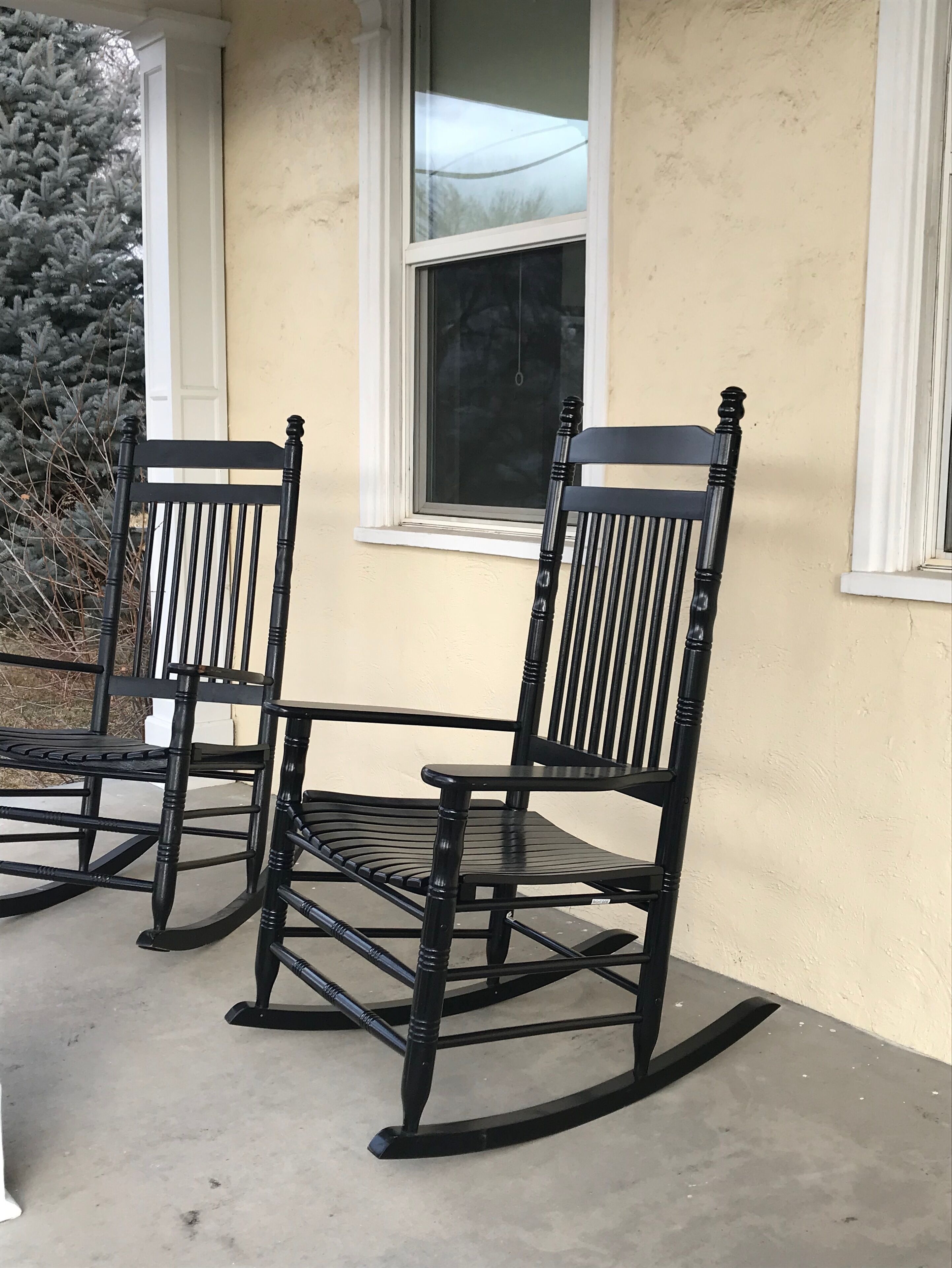 Two rocking chairs on a concrete porch with a beige wall and a window.
