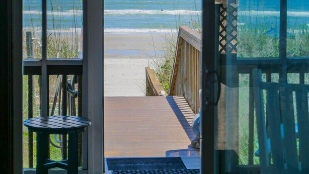 Une chambre avec vue sur la plage, donnant sur une terrasse en bois qui mène au sable, avec une petite table et une chaise.