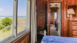 Une chambre dans un chalet en bois, avec une grande fenêtre donnant sur la plage, un lit, une commode et un miroir.