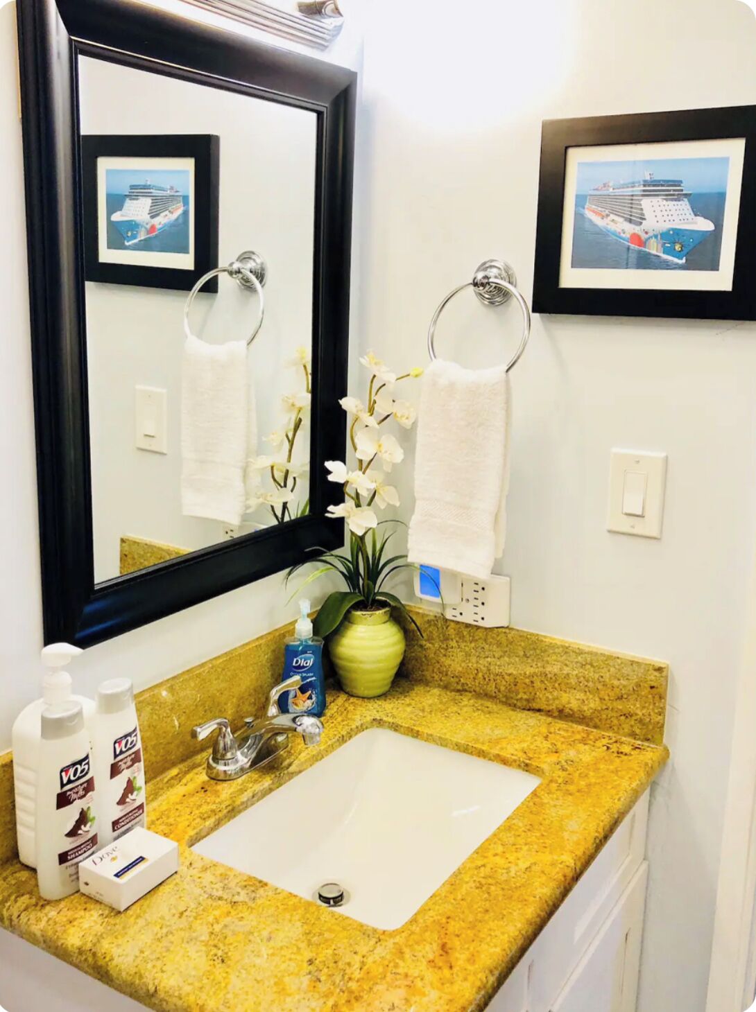 A bathroom with a granite countertop, a white sink, a mirror, two framed pictures of cruise ships, a potted orchid, and toiletries.