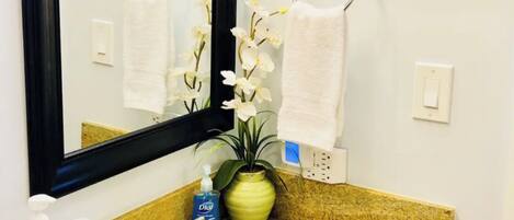 A bathroom with a granite countertop, a white sink, a mirror, two framed pictures of cruise ships, a potted orchid, and toiletries.