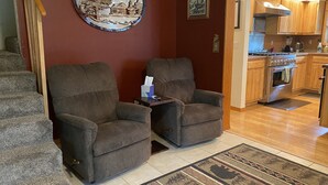 A living room with two grey armchairs, a wooden coffee table, a rug, and a kitchen visible in the background. - The Best Kenai Peninsula Base Camp. (Sterling)