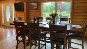 A wooden interior with a dining table set for six, surrounded by chairs, and a large window offering a view of a green outdoor area. - The Best Kenai Peninsula Base Camp. (Sterling)