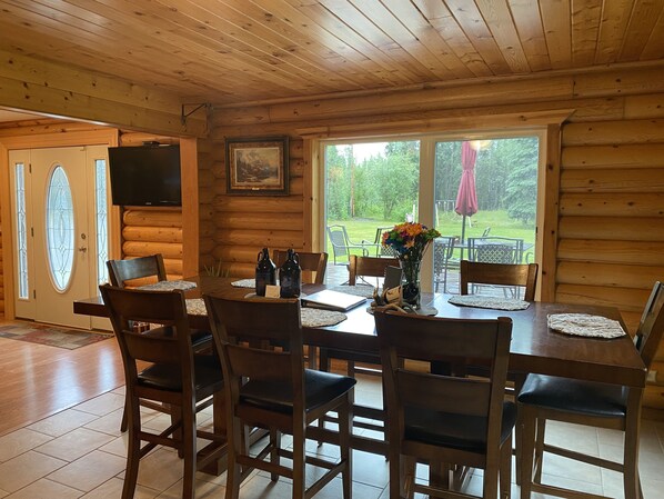 A wooden interior with a dining table set for six, surrounded by chairs, and a large window offering a view of a green outdoor area.
