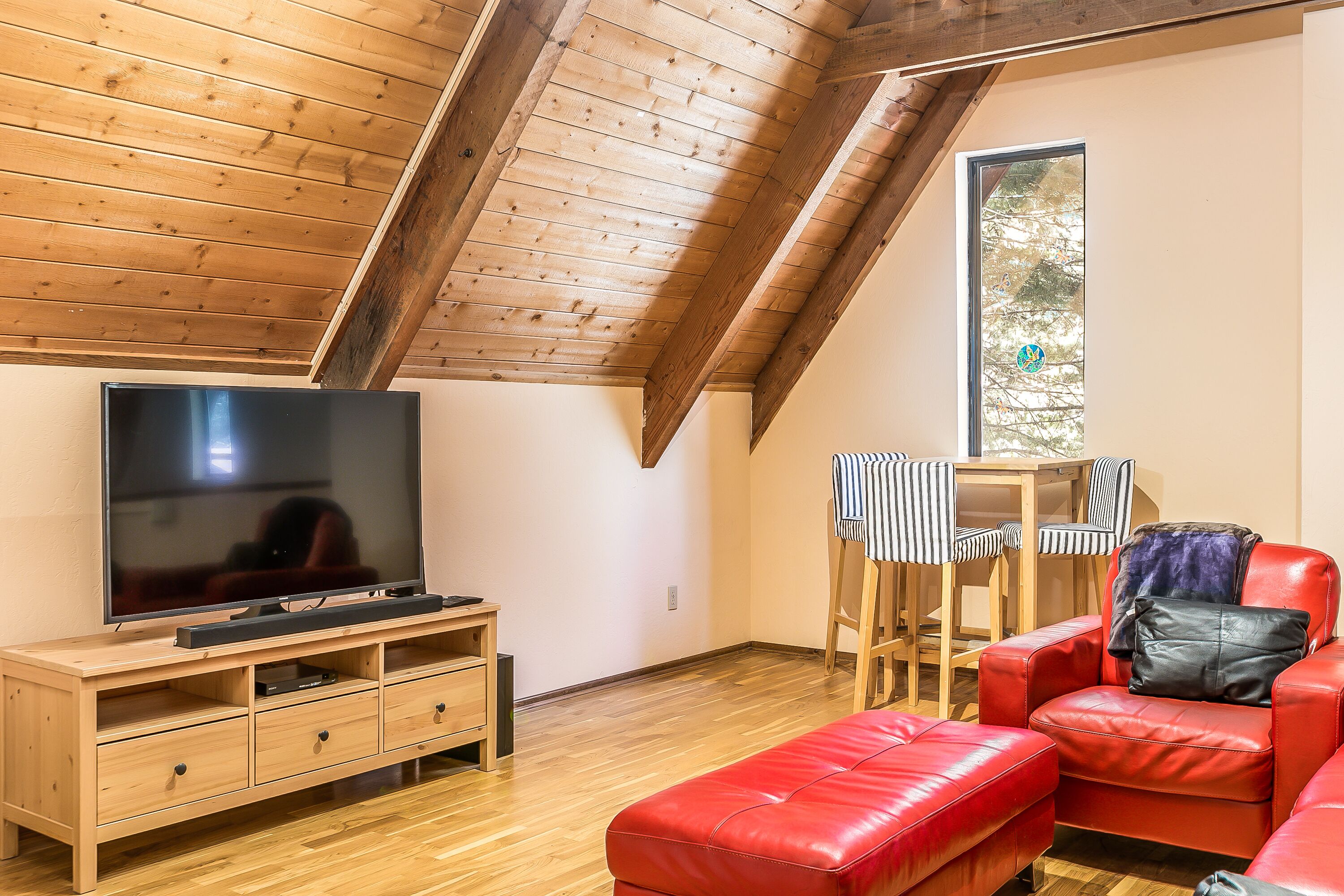 Un salon chaleureux avec un plafond en bois, un téléviseur à écran plat posé sur un meuble en bois, un canapé en cuir rouge, un fauteuil à rayures et une petite table recouverte d’une nappe à rayures.