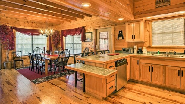 A wooden cabin interior with a dining area, kitchen, and large windows. - Lazy Bear Lodge: 6BR, Theater,Spa,Pool,Game room! (Pigeon Forge)
